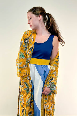Woman wearing a long yellow floral kimono over a blue top and white skirt against a plain background