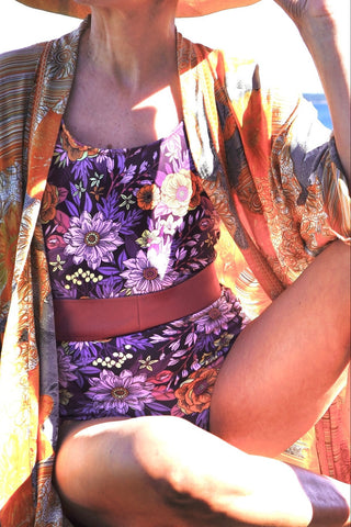 A close-up of a person wearing a purple floral one-piece swimsuit with a wide pink belt, layered under a patterned orange and gold kimono-style cover-up.